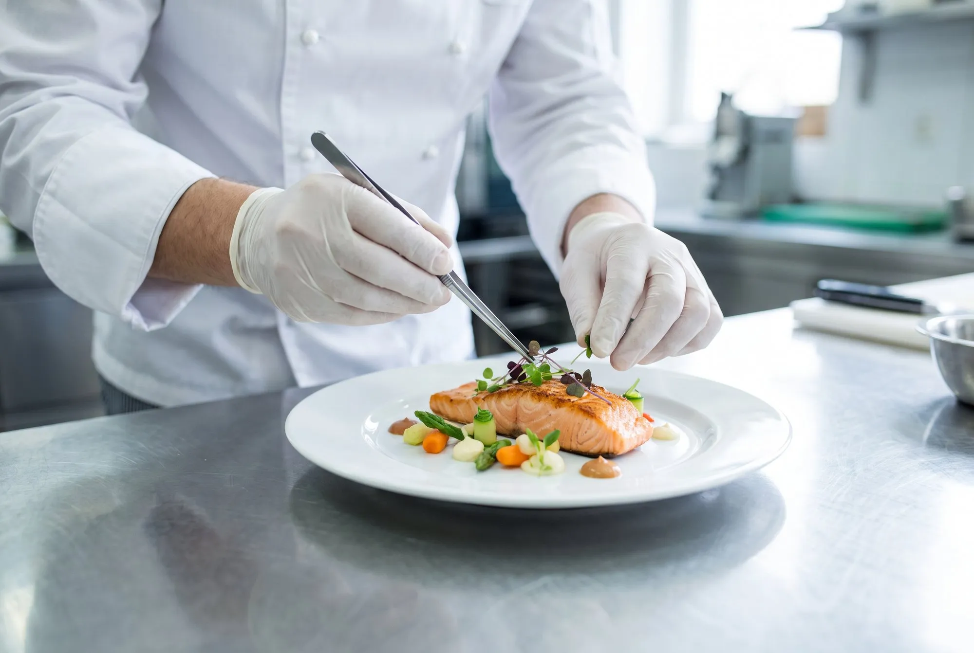 A chef plating a dish with tweezers, seen over the shoulder