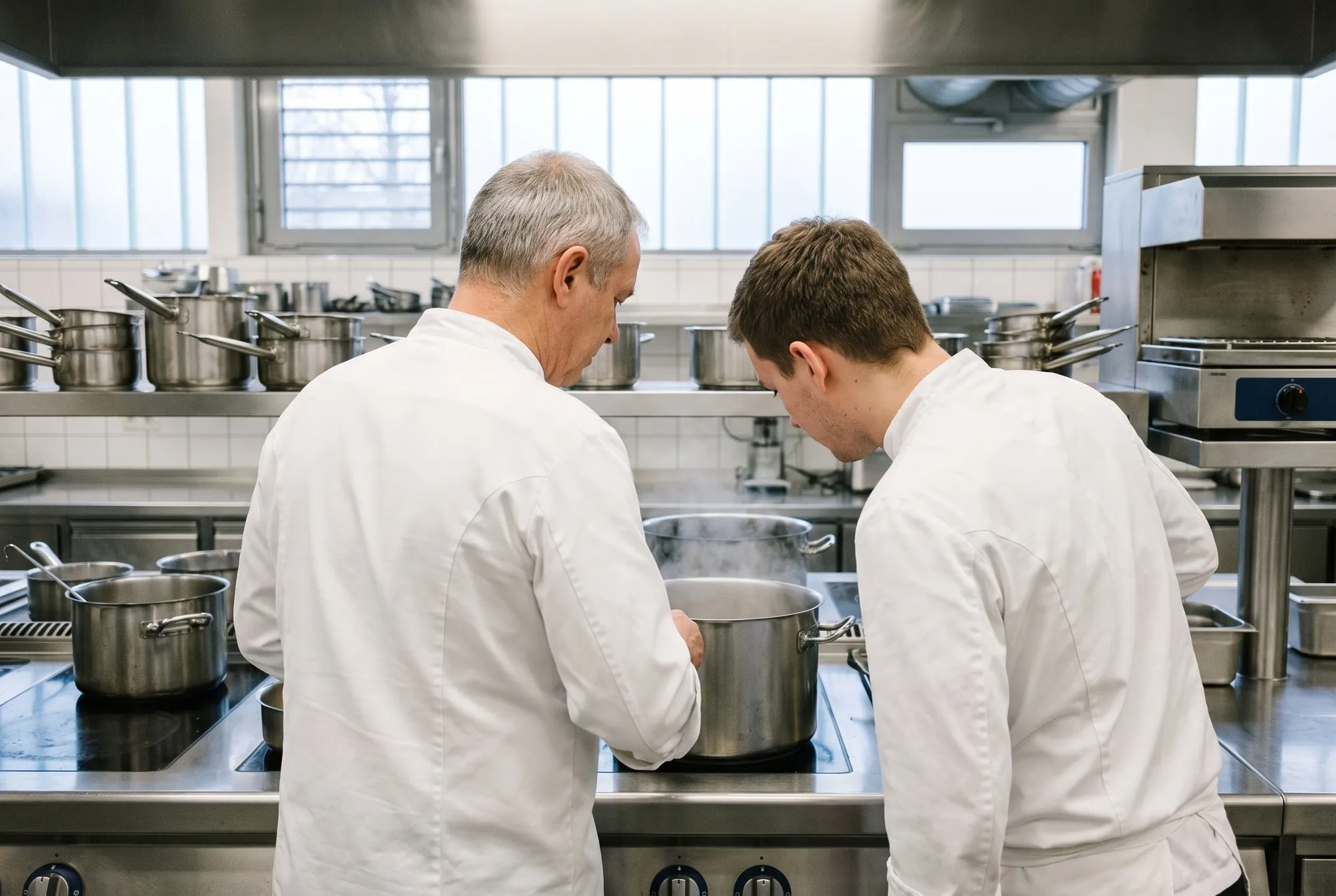 Two chefs seen from behind at the stove, one explaining a step